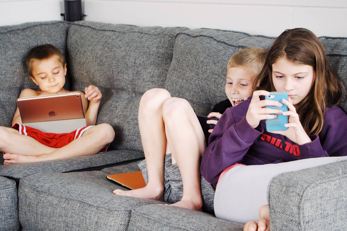 three kids on a couch looking at phones and tablets