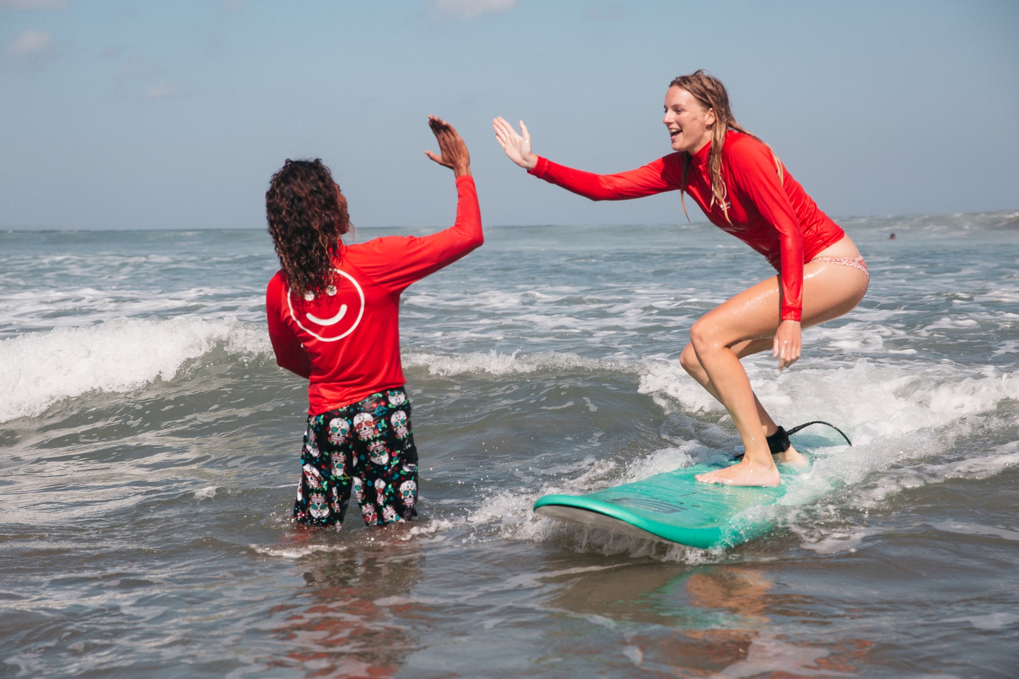 A surfer with black skin and long brown dreads and a surfer with white skin and long brown hair both in red rash guard UPF shirts high fiving in the waves