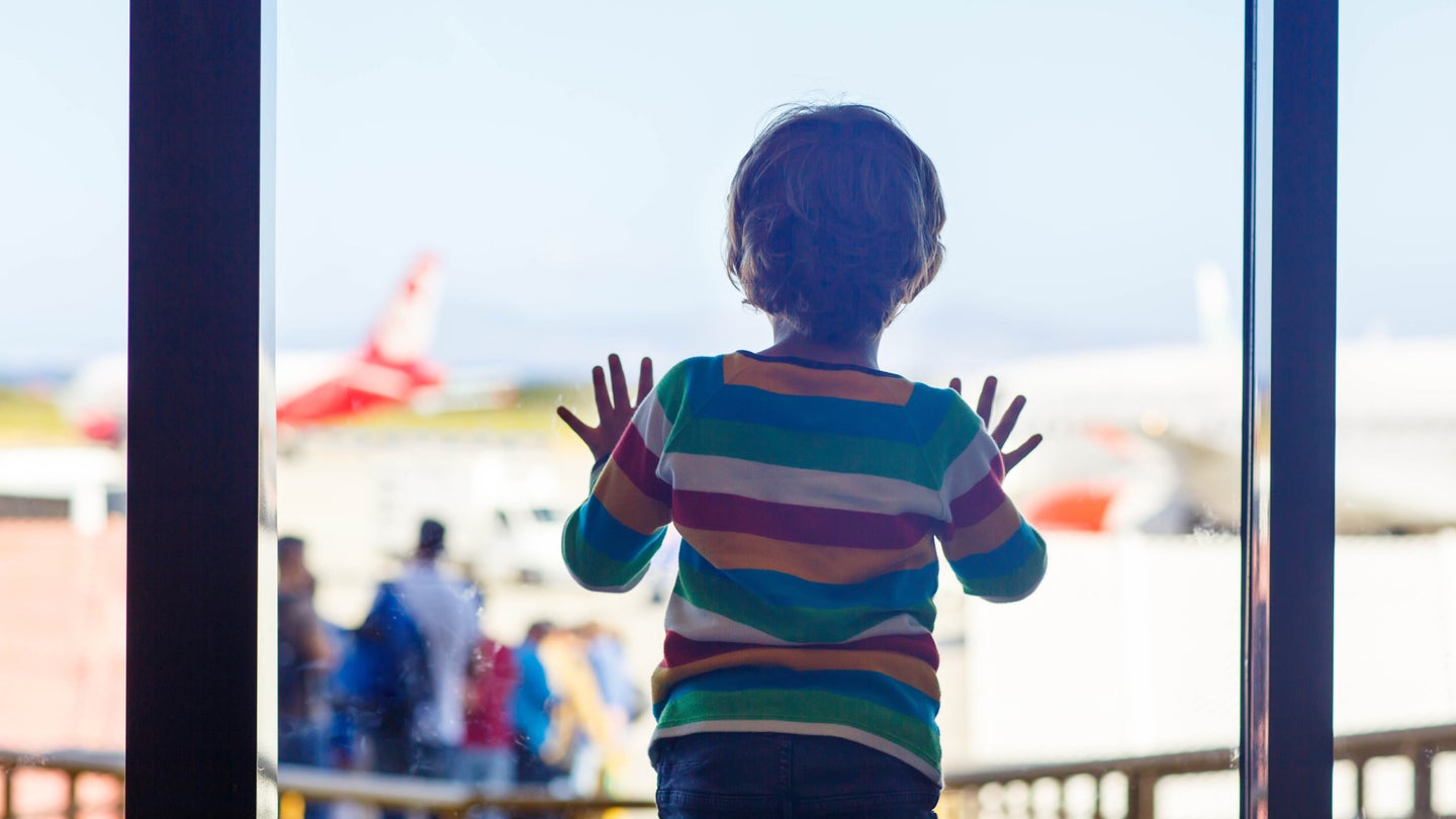 A small child standing at a large glass window in an airport, looking out at the planes on the tarmac.