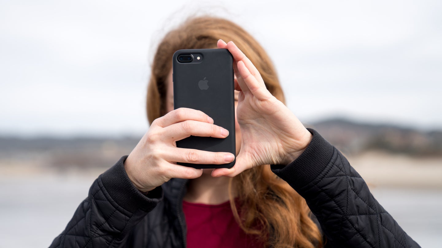 A red-haired woman facing the camera, using her black iPhone to take a photo, with the phone covering her face.