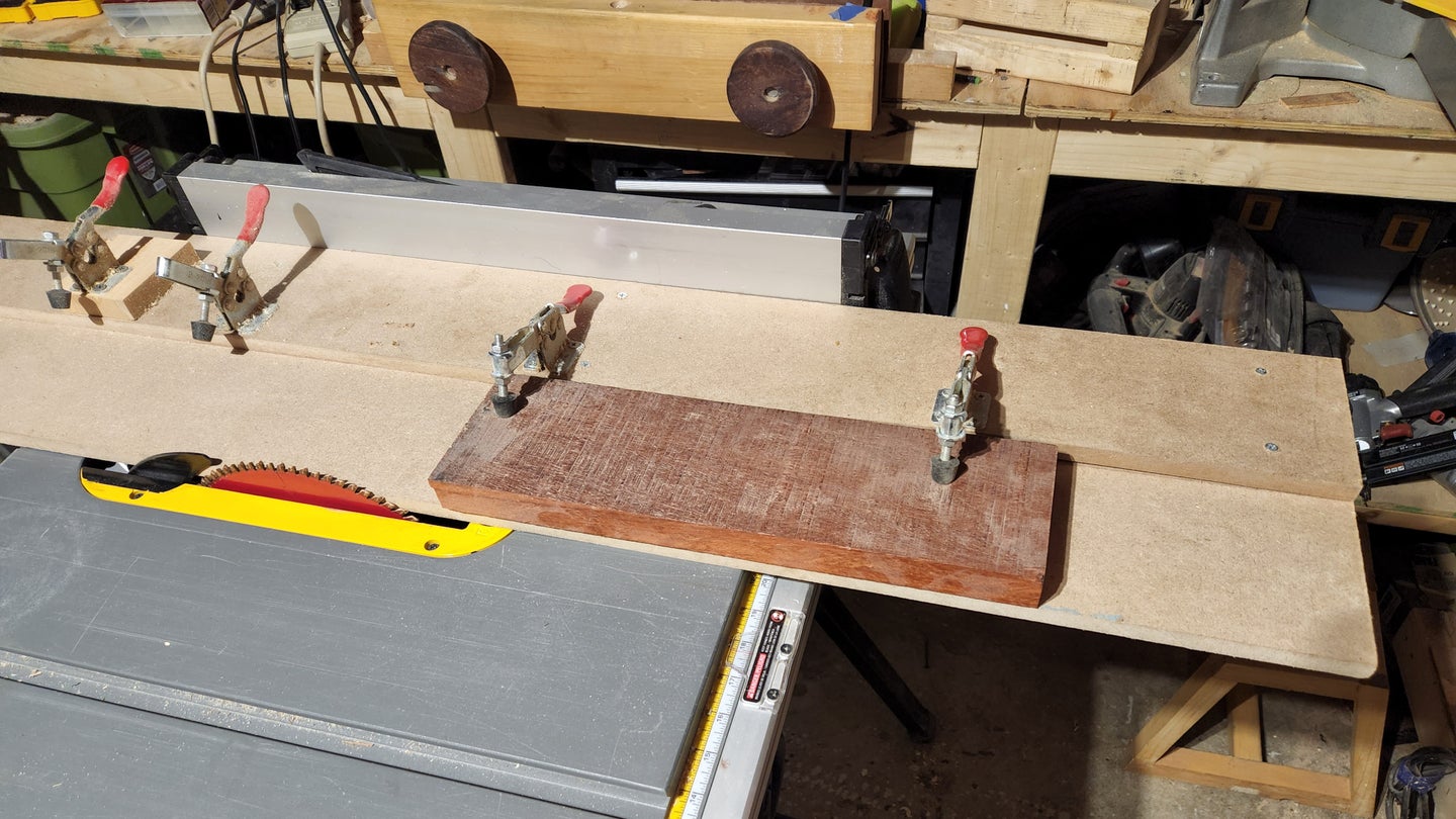 A DIY jointer sled holding a small piece of wood in place atop a table saw in a woodworking shop.