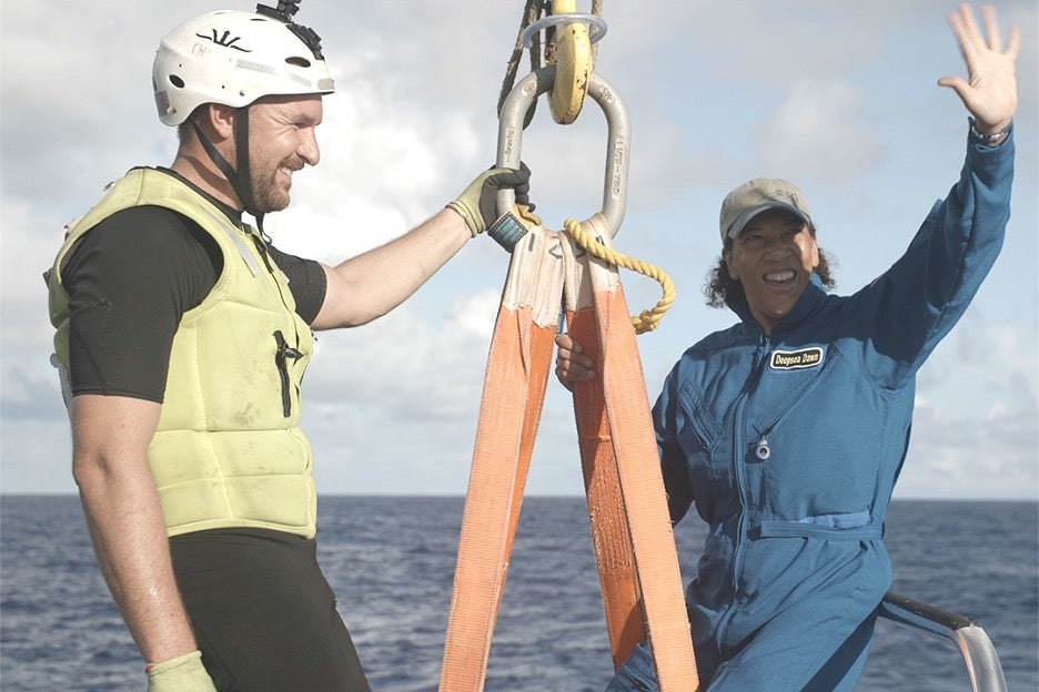 A black woman in diving gear and a hat excitedly waves next to a man holding a rope in Substeady.