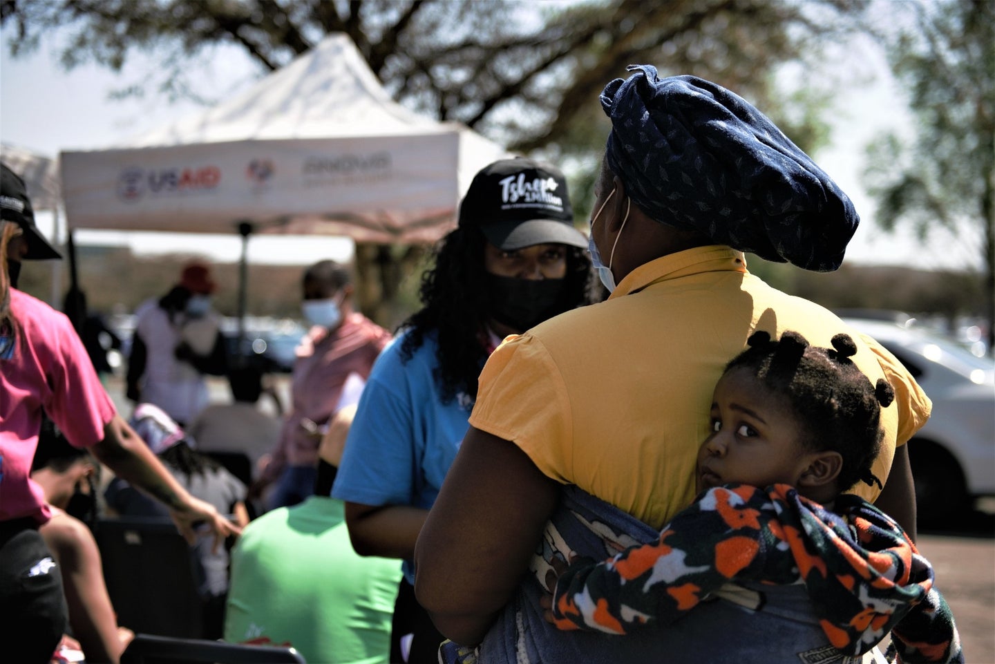 Mother in yellow shirt and blue head wrap with child strapped to back at a COVID vaccine clinic in South Africa