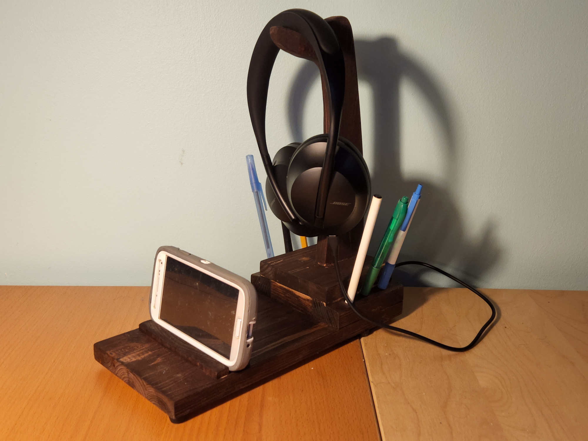 A homemade wooden DIY headphone stand on a lighter-colored wooden table, complete with a phone holder, a USB charging port, and slots for pens and pencils.