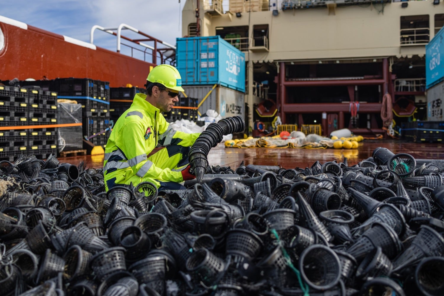 a man in a yellow hazard working suit stacks black plastic cones on a ship