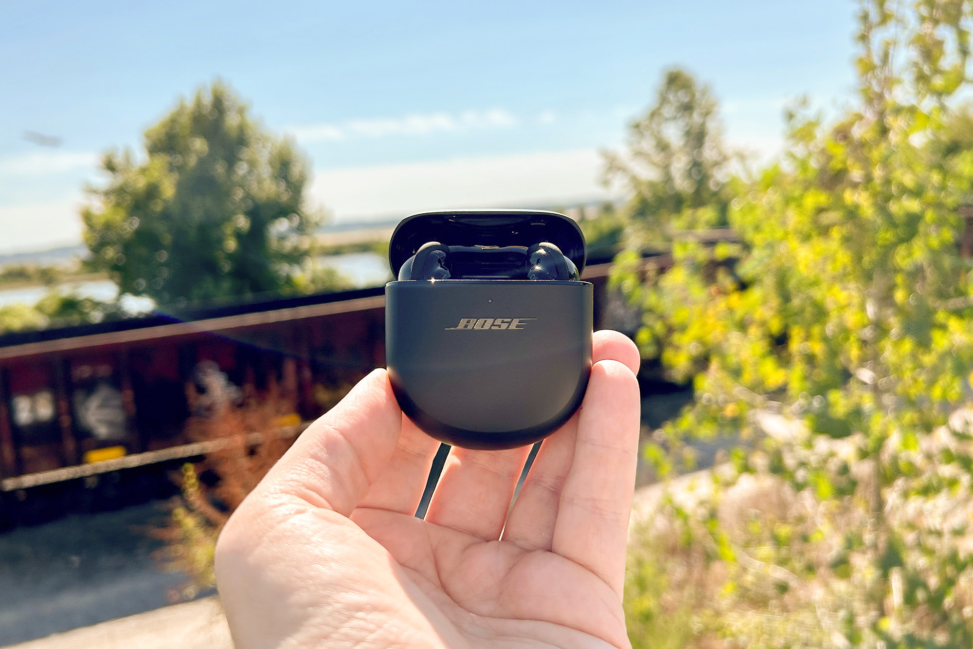 Bose QuietComfort Earbuds II outside with a train and plane in the background