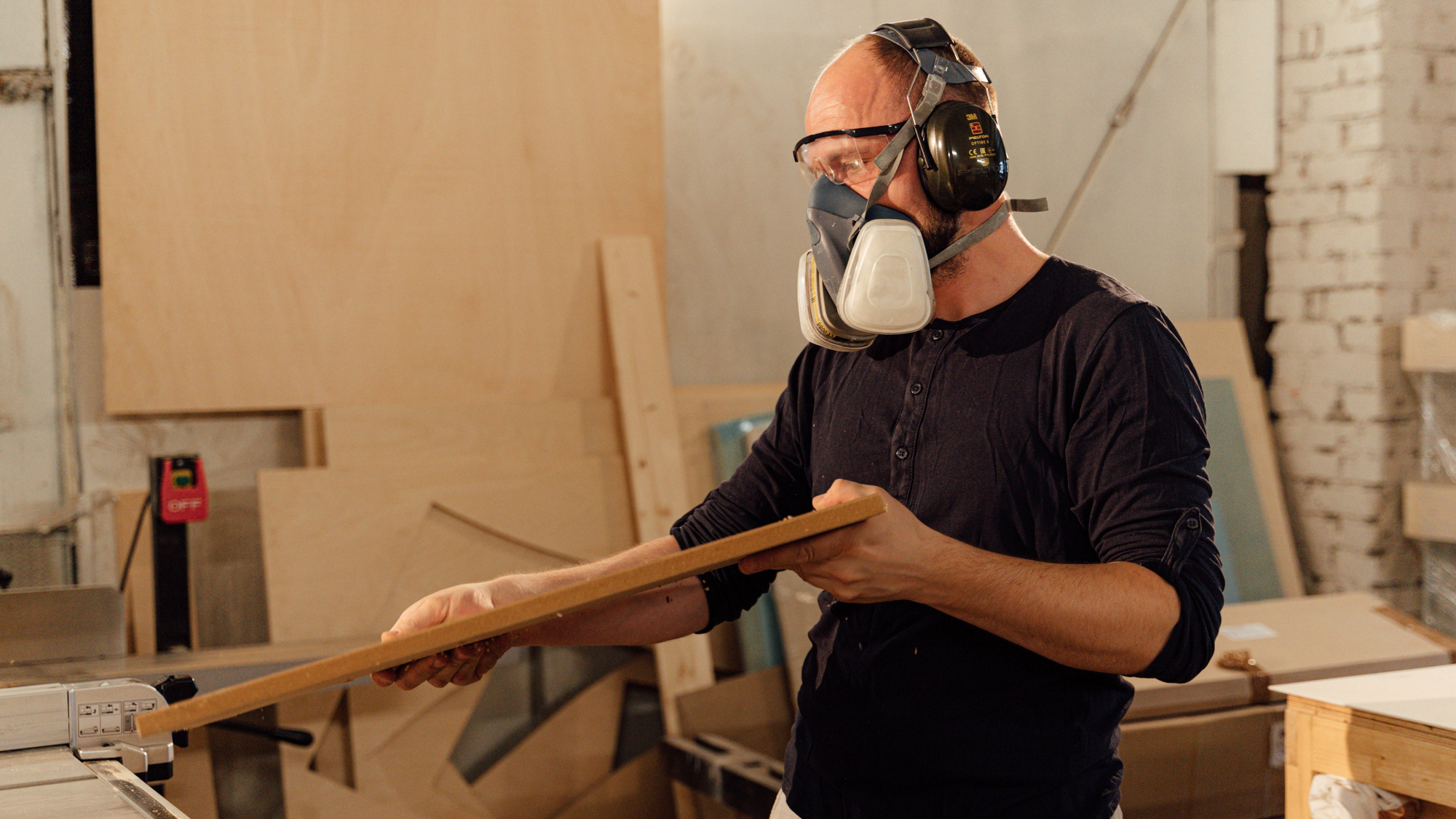 A bald man wearing a dust respirator and earmuffs working in a woodshop and milling his own wood to make it straight.