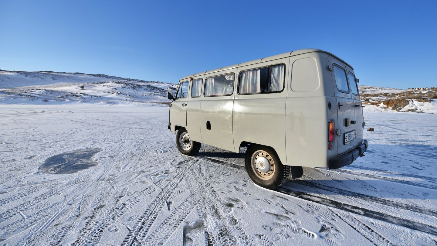 A white camper van with curtains parked on a flat, open expanse of snow under blue skies, near some low mountains or hills.