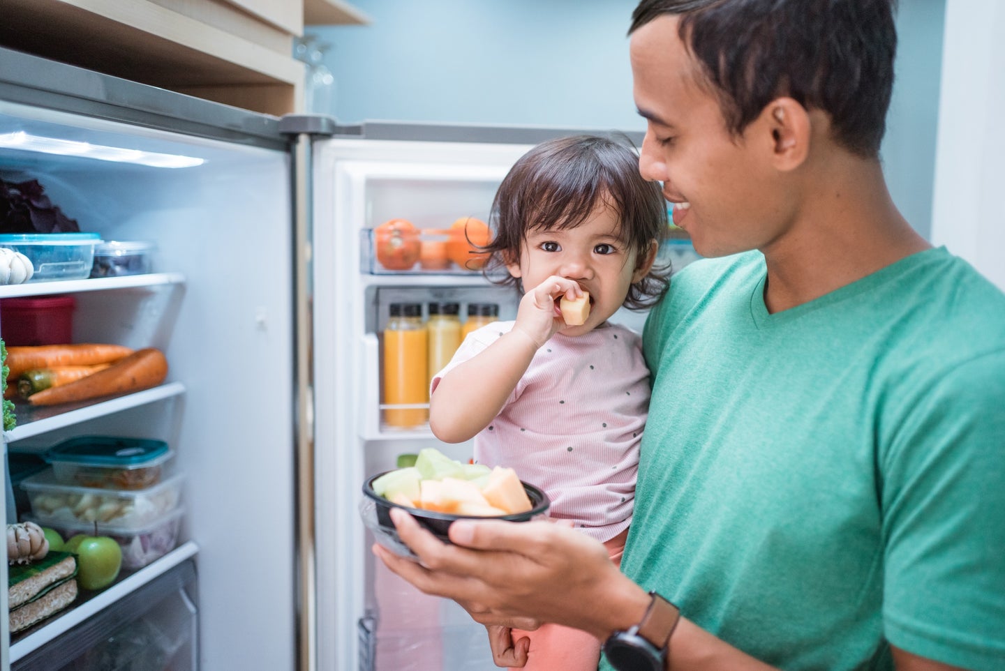 Brown-skinned parent in green short and baby in pink onesie eating fruit in front of an open fridge, which is full of HFCs