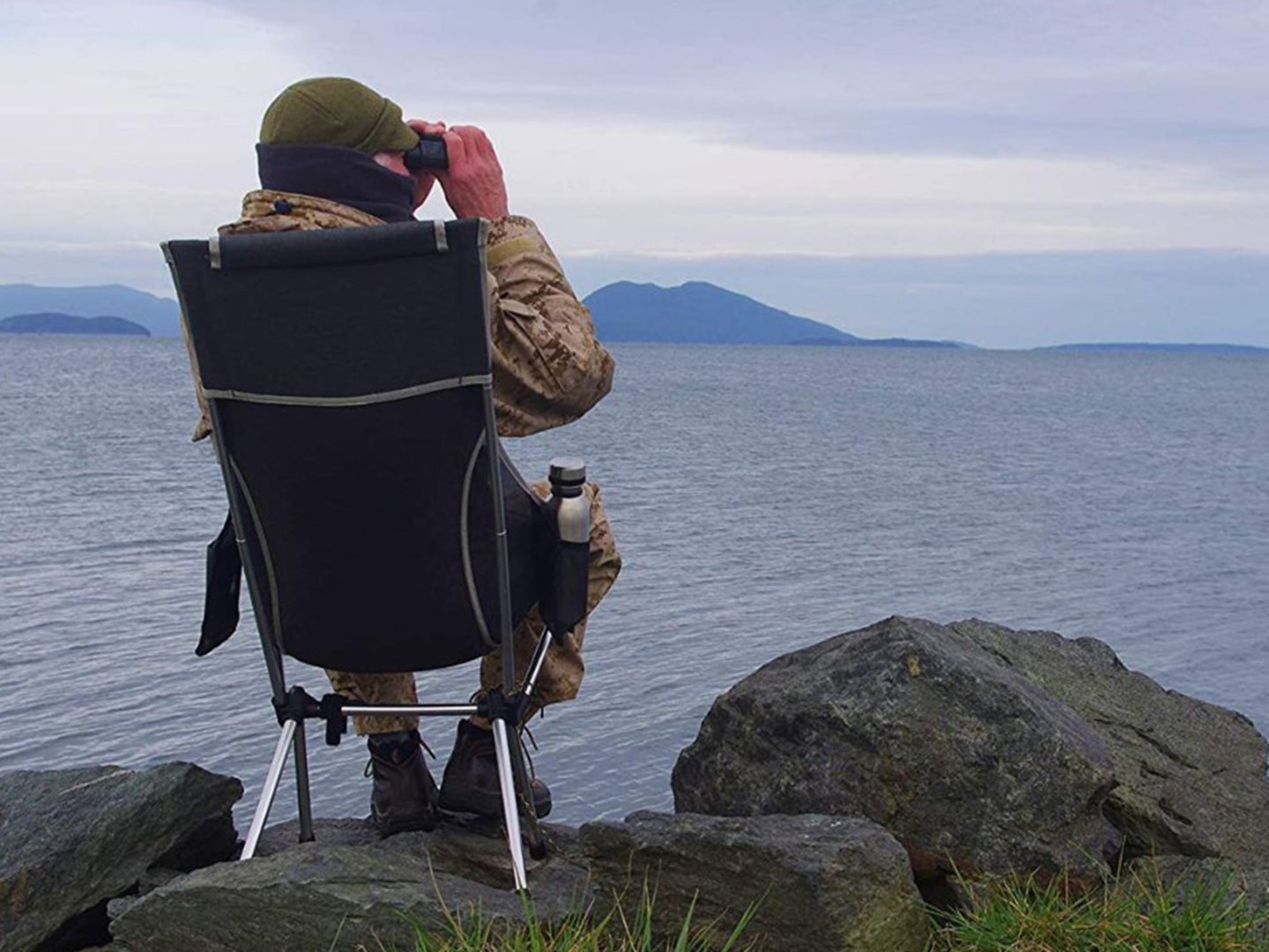 A person sits on a folding camping chair and looks out into a lake with binoculars