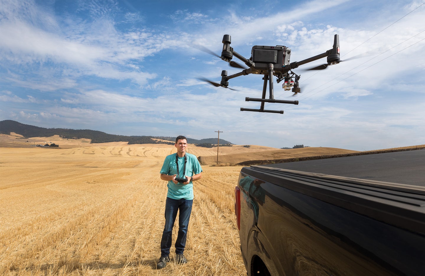 A man standing in a field lands a drone in the back of a pickup truck.