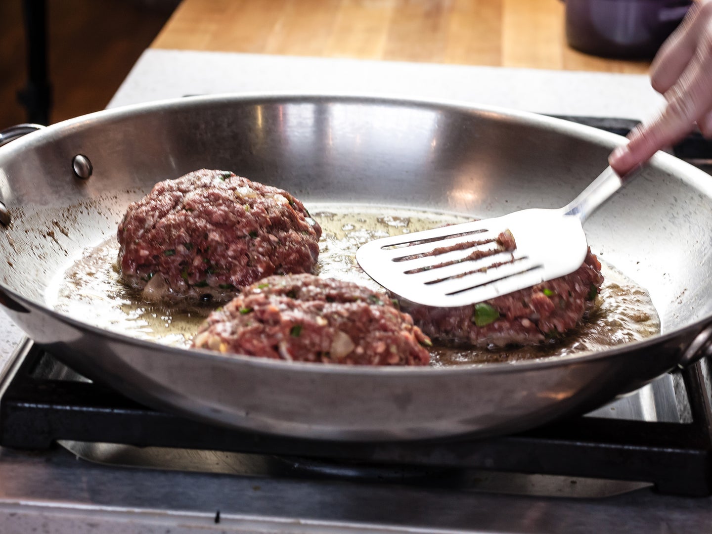 Three burgers getting cooked on a stainless steel pan while a hand with a spatula works with them