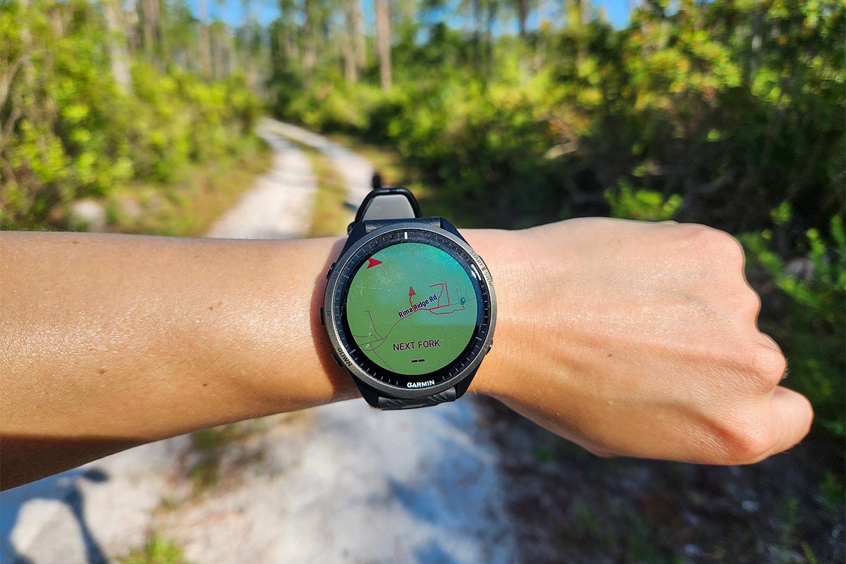 A woman wearing a Garmin Forerunner 965 with a green face on her wrist against the background of a hiking trail.