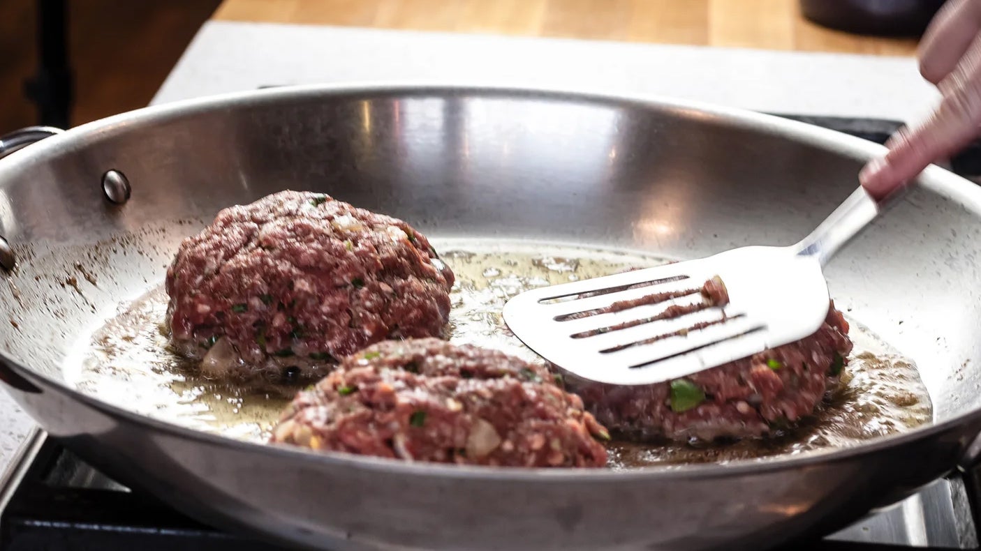 Three burgers getting cooked on a stainless steel pan while a hand with a spatula works with them