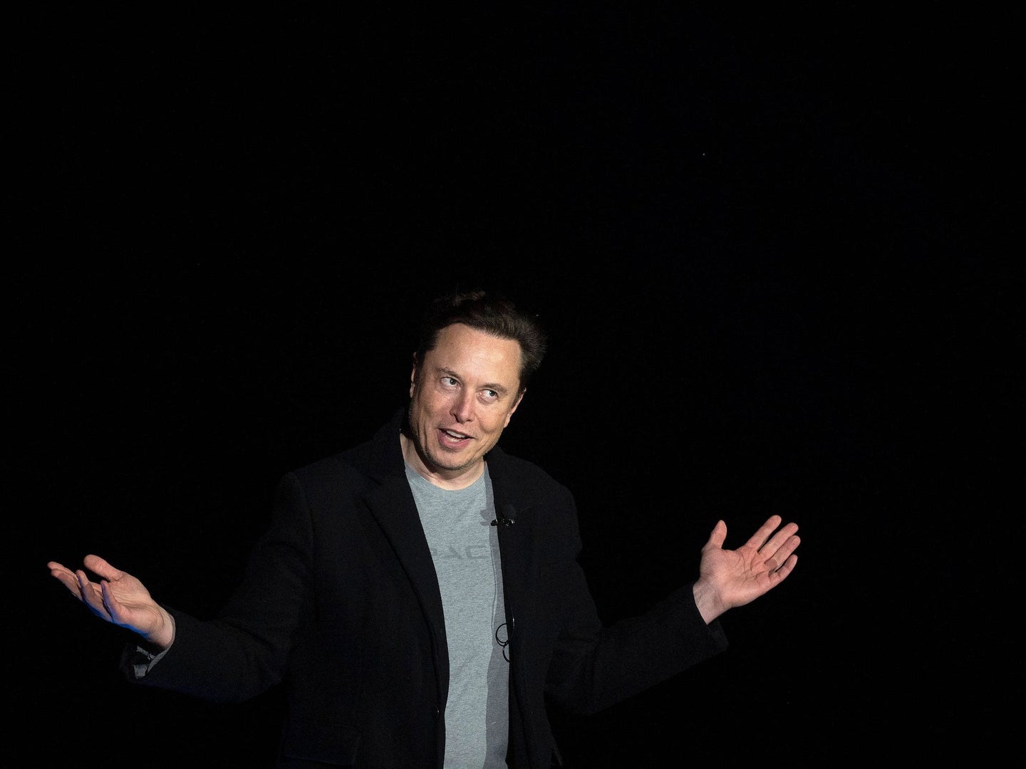 Elon Musk gesturing with both hands up against black background