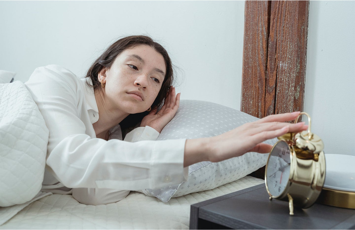 A woman in a long sleeve white shirt reaches from bed to turn off her alarm clock.