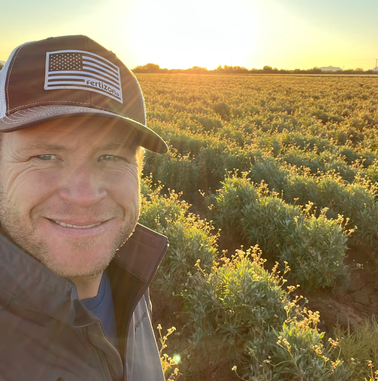 a farmer in a baseball cap takes a selfie in front of a farm of guayule