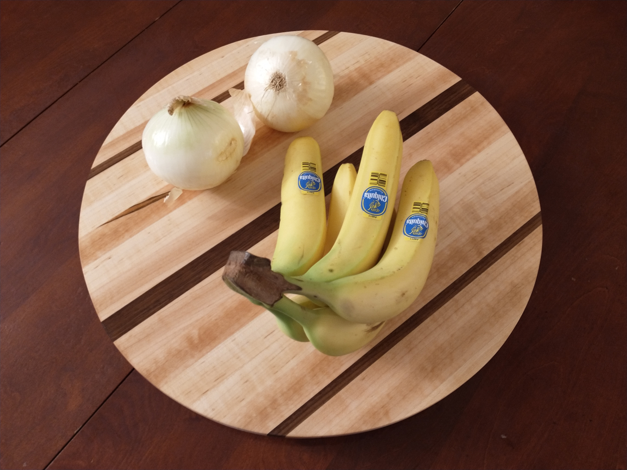 A DIY Lazy Susan for a tabletop, on top of a dark wooden table. The Lazy Susan has a bunch of bananas and two white onions on it.