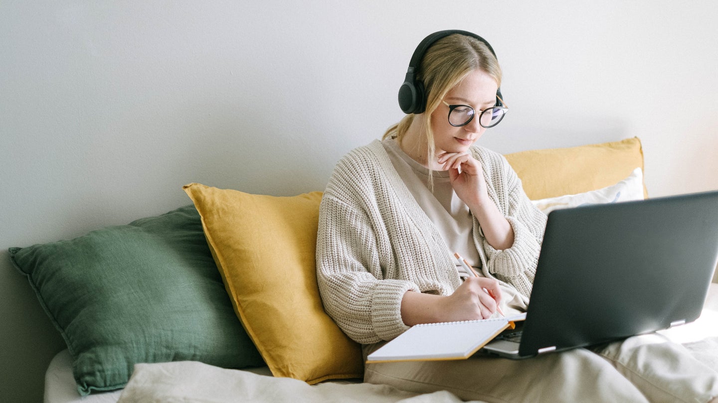 A blonde woman wearing a white sweater and headphones while sitting on a couch or bed, taking notes while in front of a laptop computer.