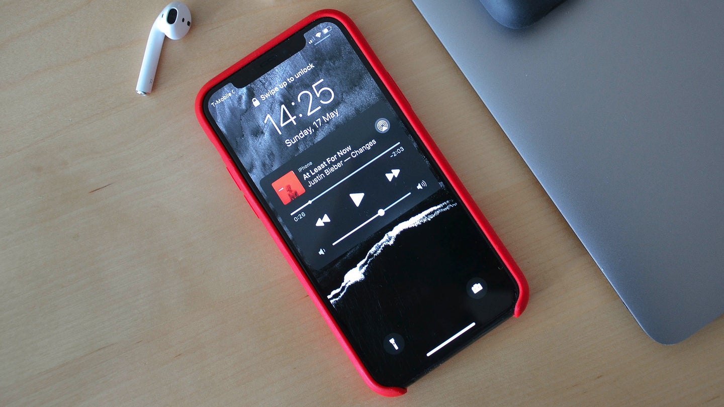 An iPhone on a desk by a laptop and showing Apple Music on its lock screen