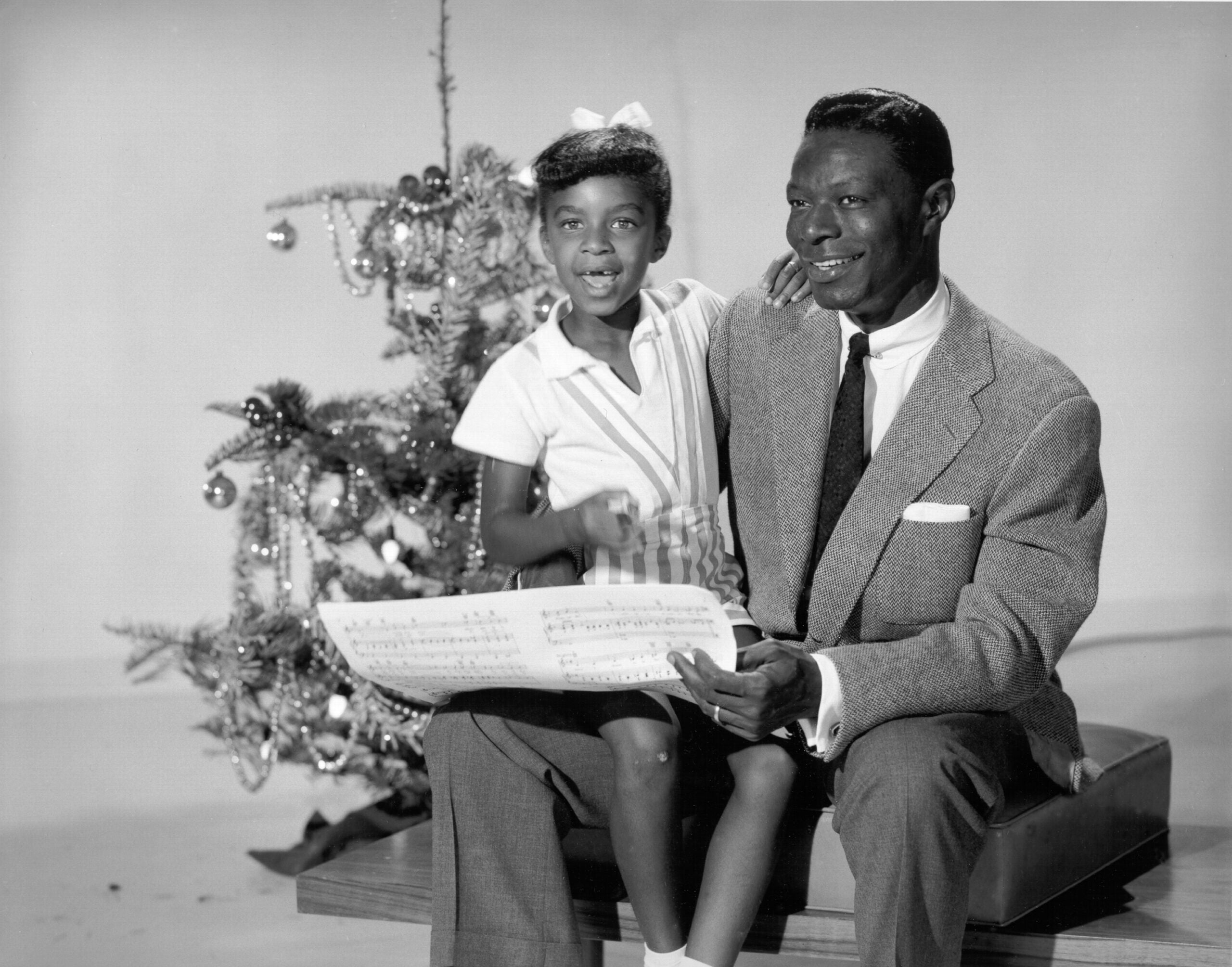 Nat King Cole and his daughter Natalie Cole in front of a Christmas tree singing Christmas songs. Black and white photo.