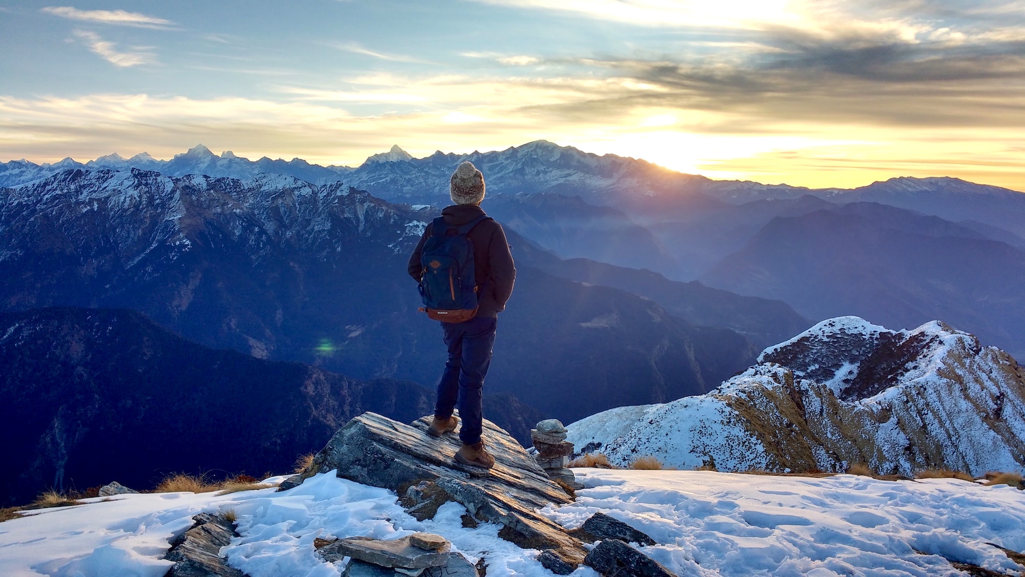 a person stands on a snow covered mountain top looking out a vista of rocky mountains as the sun peeks above the horizon