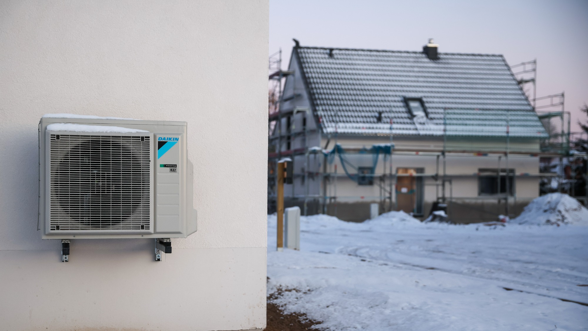 An air-source heat pump hangs on a house wall in snow and ice in a newly developing single-family housing area.