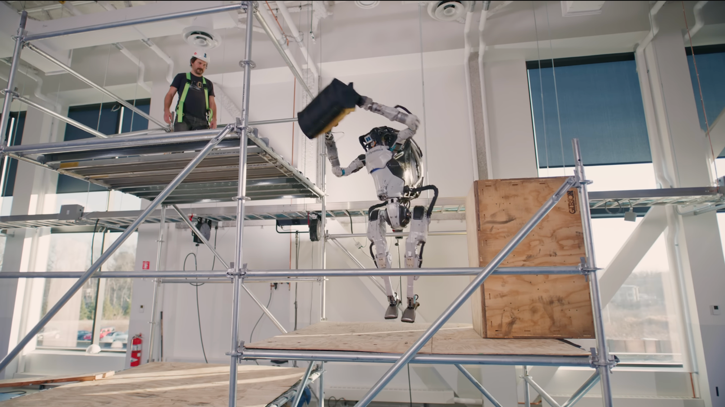 Boston Dynamics Atlas bipedal robot tossing toolbag up to human on construction scaffolding in warehouse