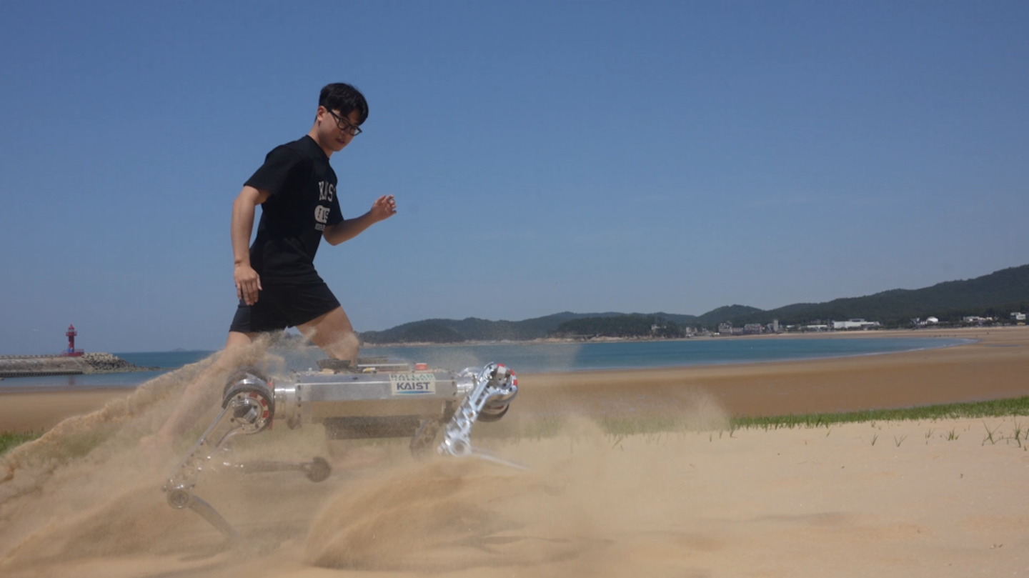 Four legged robot running alongside man on sandy beach