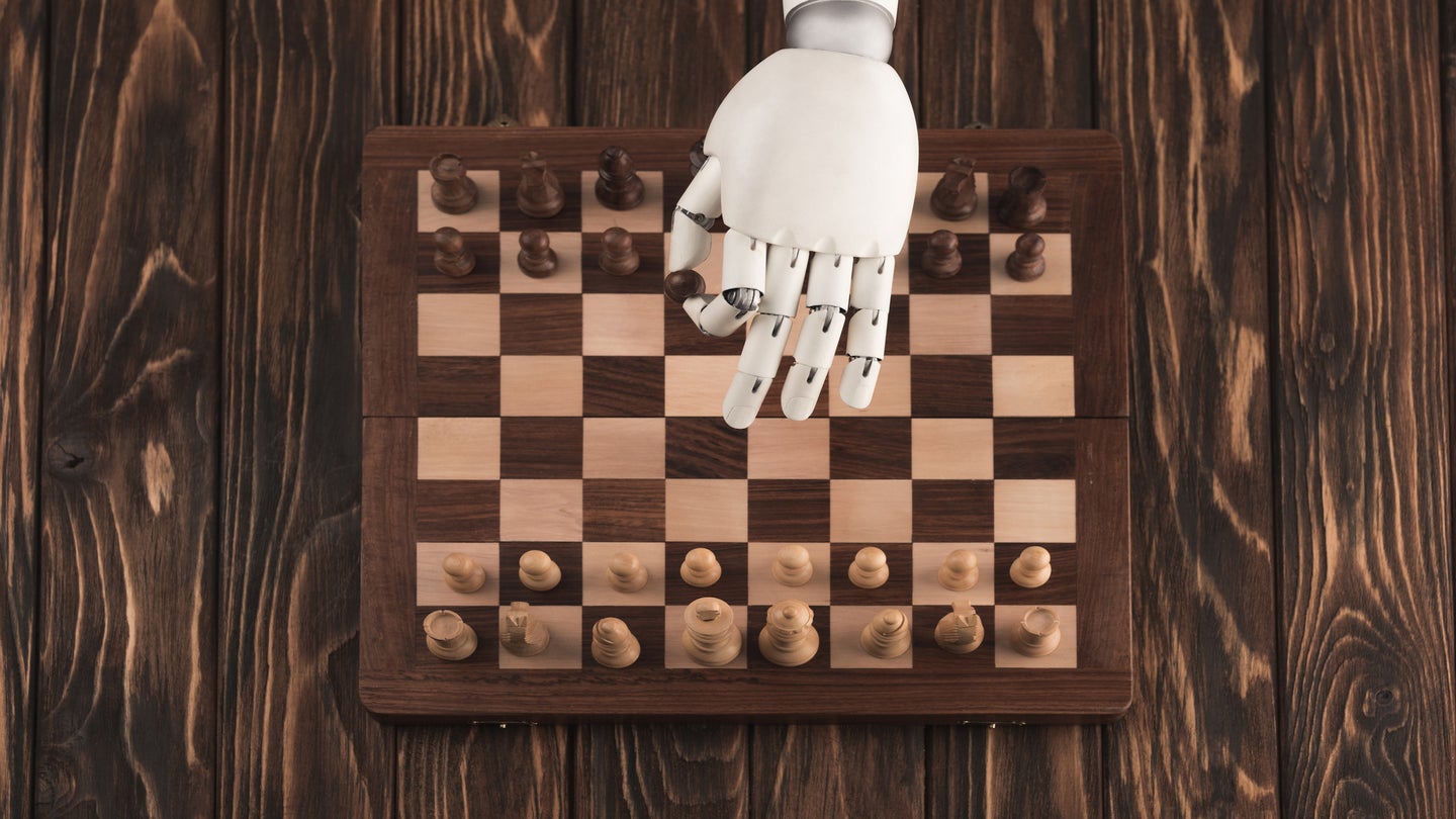 A white robotic hand moving a black pawn as the opening move of a chess game played atop a dark wooden table.