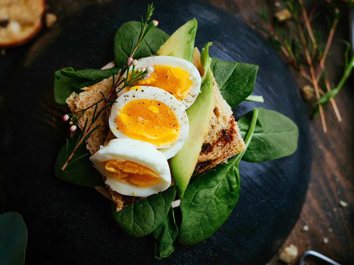 A handful of spinach is topped with a slice of bread, avocado, and medium-boiled eggs on a wooden table.