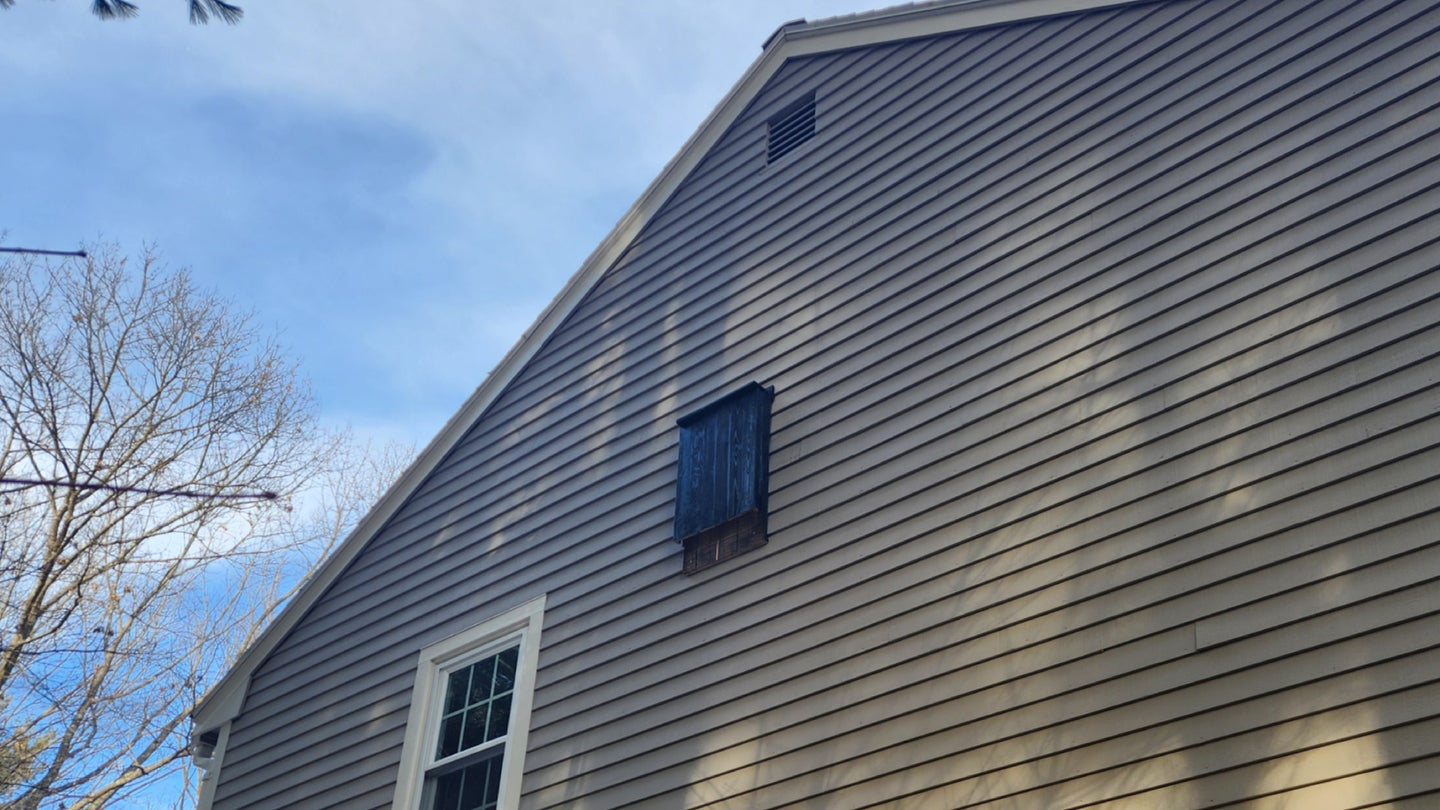 A dark wooden bat house high up on the side of a gray two-story house.