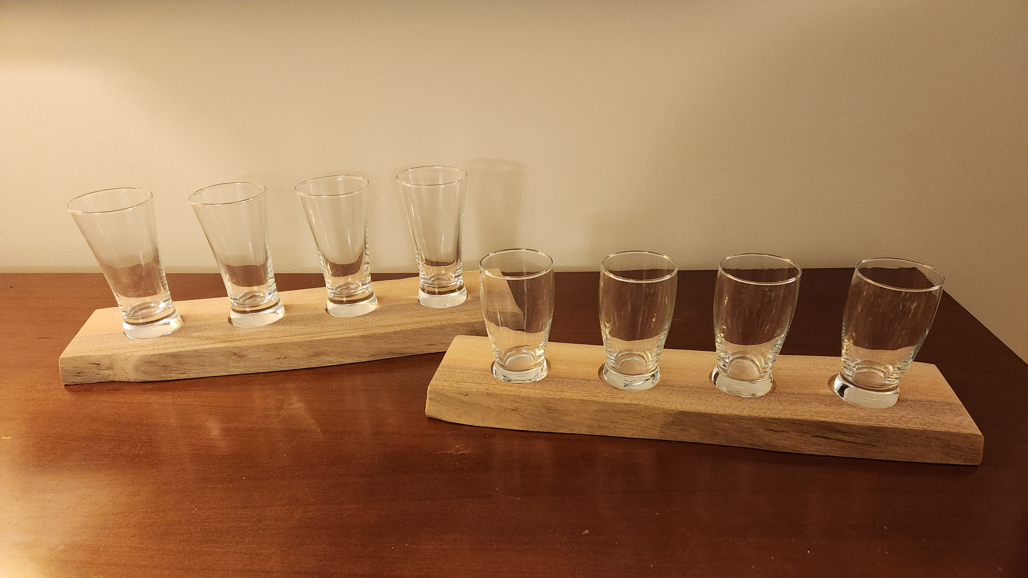 Two light-colored live edge DIY beer flight boards with four glasses each, on a darker wood surface in front of a beige wall.