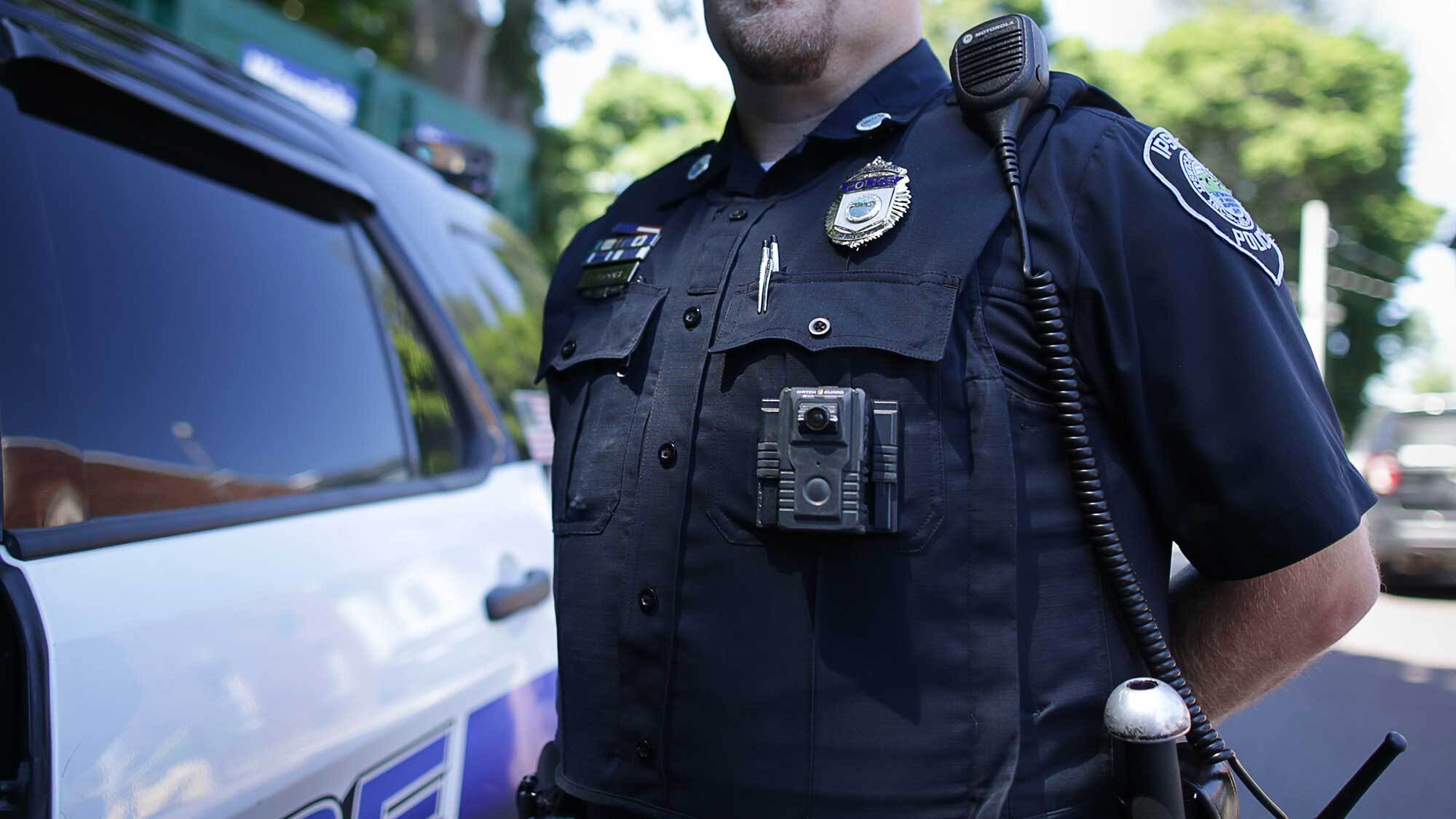 Chest of police officer in uniform wearing body camera next to police car.