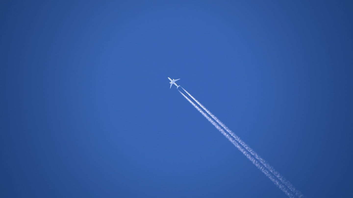 A plane flying high above the ground, drawing white contrails across a blue sky.