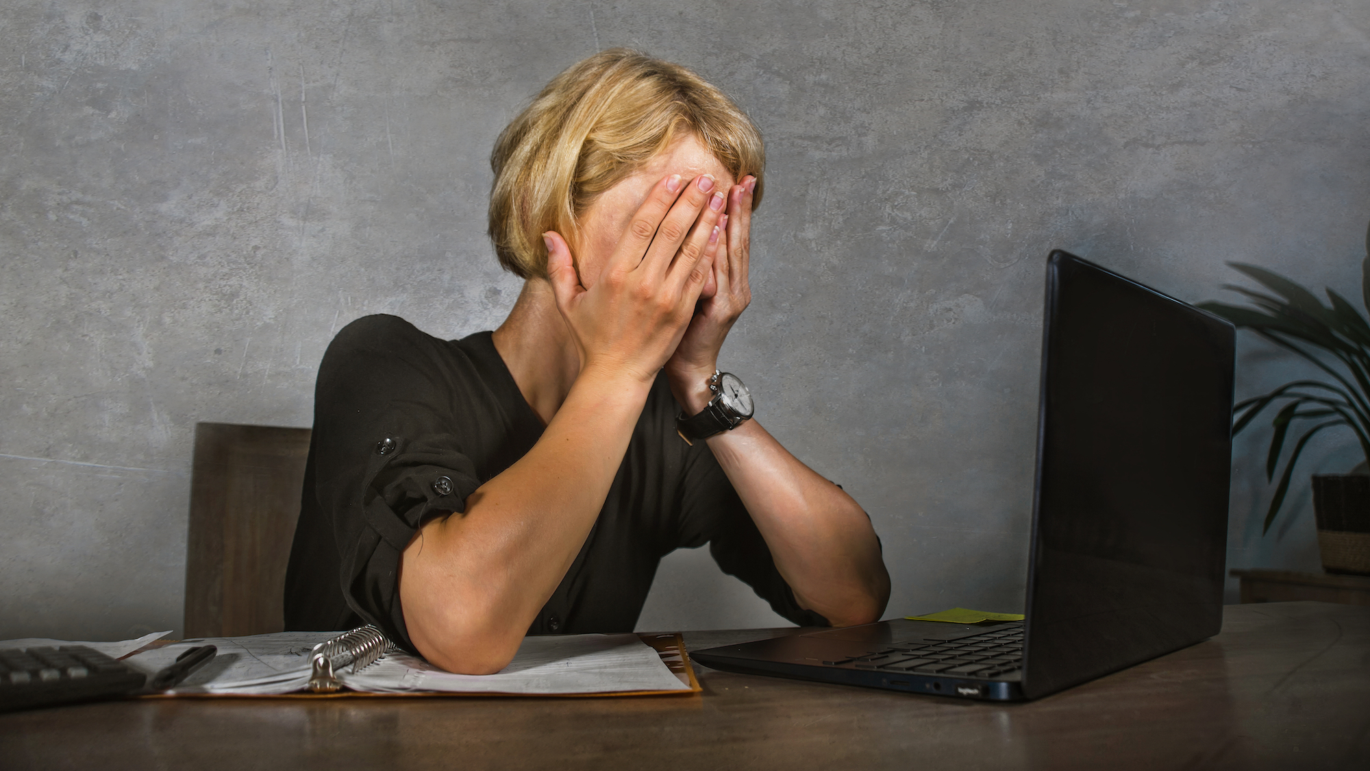 Woman covering face with hands in frustration while sitting in front of laptop on an office desk.