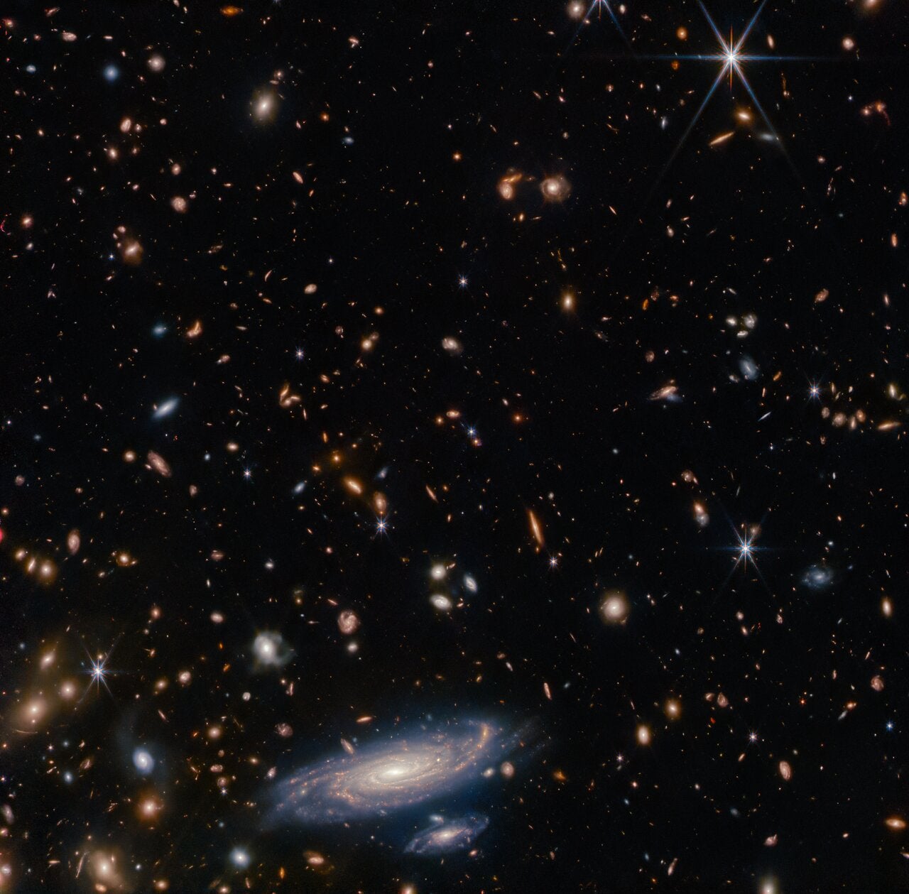 This image displays a wider view of the field of stars and galaxies surrounding the spiral galaxy LEDA 2046648. Webb’s NIRCam instrument has picked out a profusion of smaller, more distant galaxies and bright stars around this galaxy, demonstrating the telescope’s impressive resolution in infrared wavelengths. Calibration images such as this one were critical to verify the telescope’s capabilities as it was prepared for science operations, and this one doesn’t disappoint.  [Image description: Many stars and galaxies lie on a dark background, in a variety of colours but mostly shades of orange. Some galaxies are large enough to make out spiral arms. Along the bottom of the frame is a large, detailed spiral galaxy seen at an oblique angle, with another galaxy about one-quarter the size just beneath it. Both have a brightly glowing core, and areas of star formation which light up their spiral arms.]