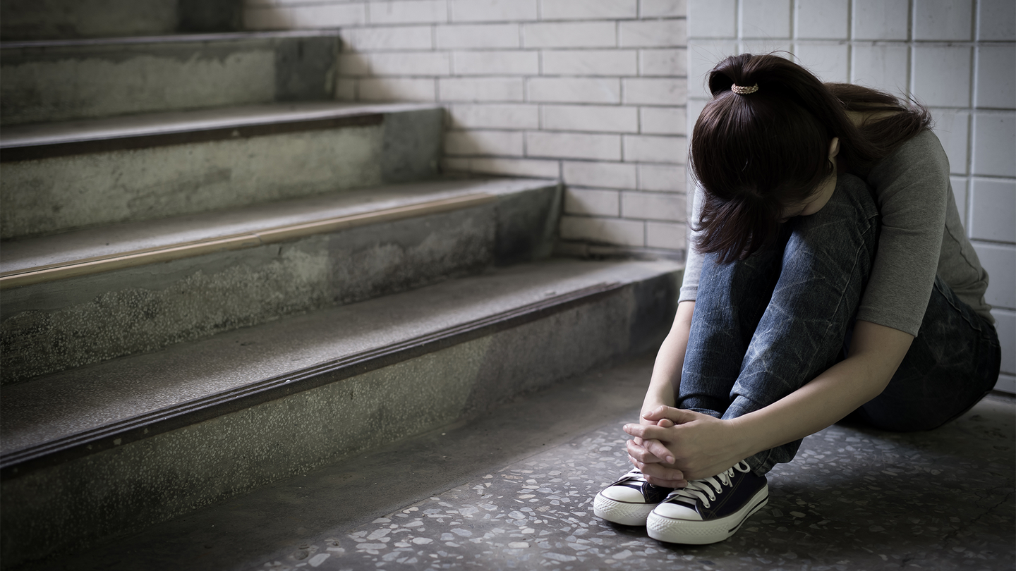 A young woman sits below a stairwell with her head down in sadness.