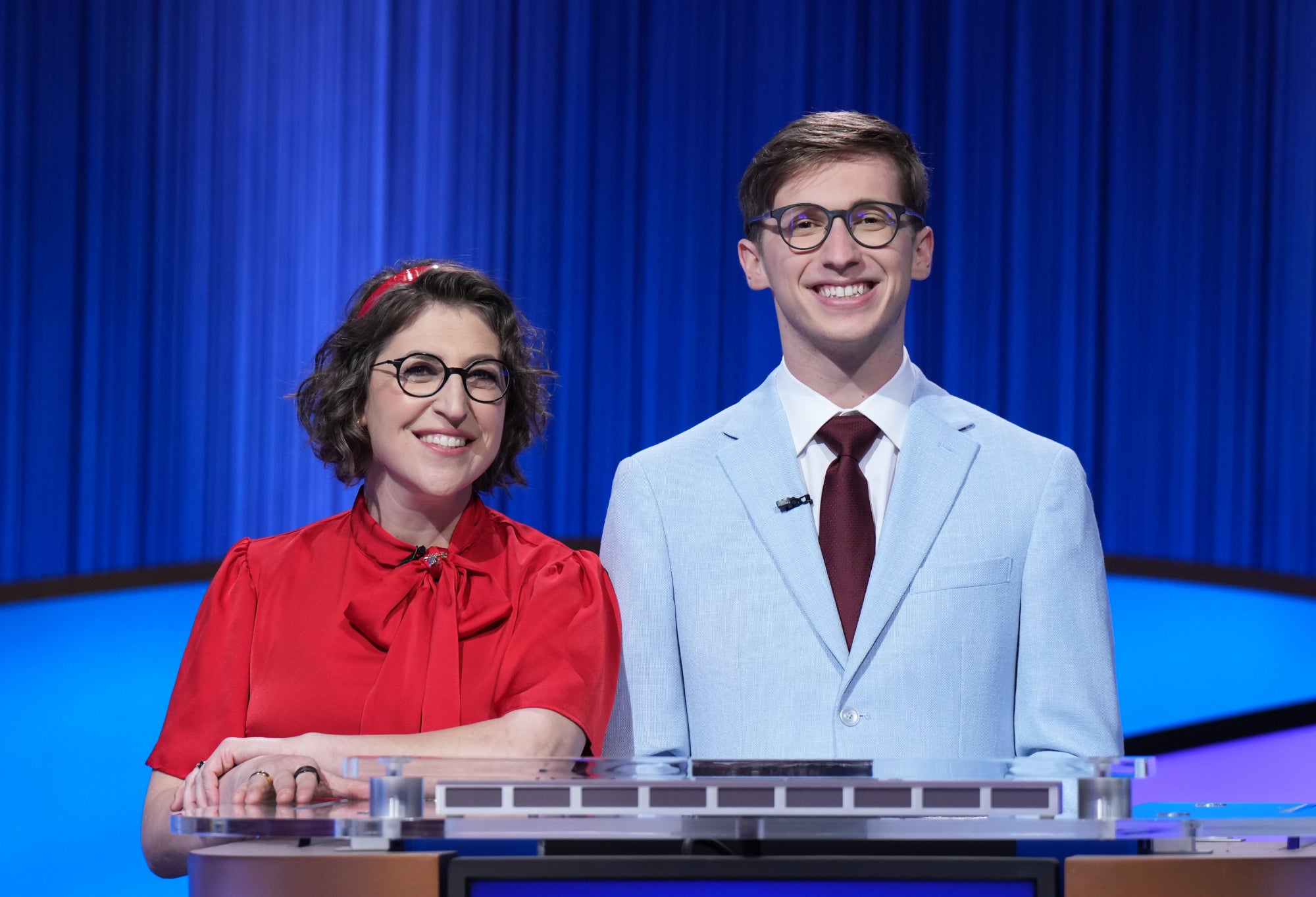 Jeopardy! host Mayim Bialik wearing red, on the left, standing next to this story's author, Jack Izzo, wearing light blue, on the right, both behind a Jeopardy! podium.