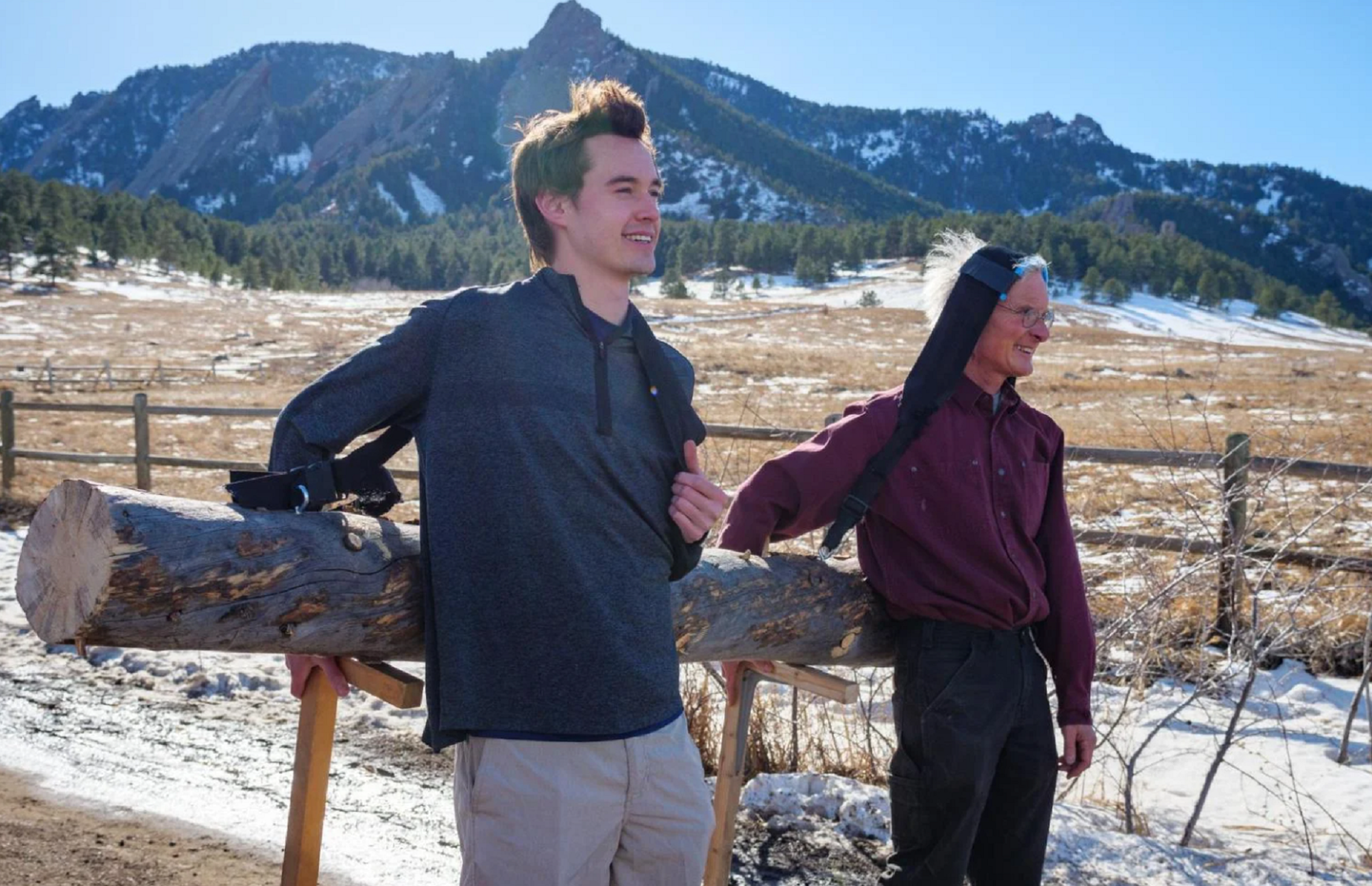 Two University of Colorado Boulder scientists in jackets carrying a log with a tumpstrap and their heads