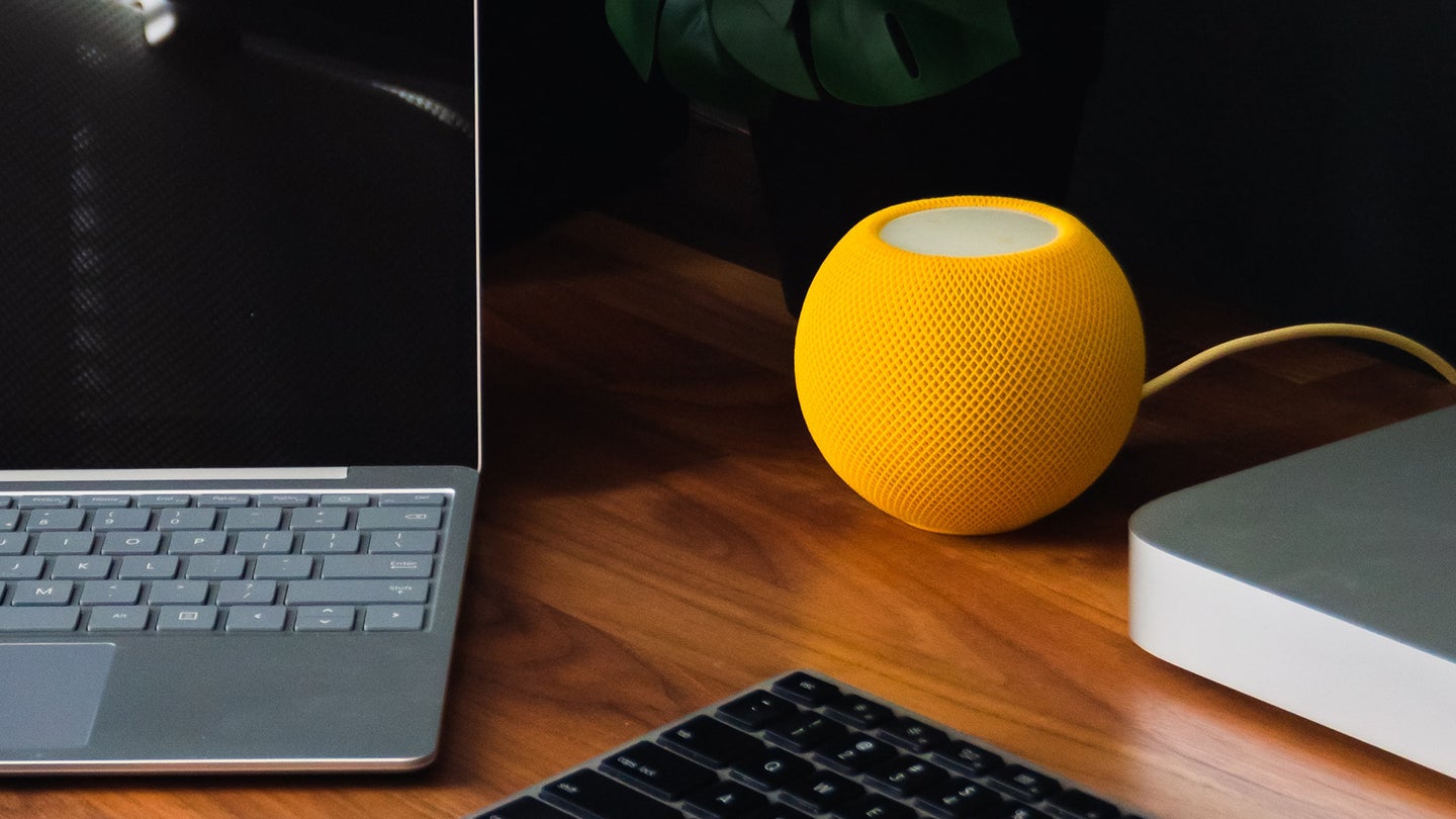An orange HomePod Mini device sits on a wooden desk right next to a laptop, a keyboard, and an external hard drive.