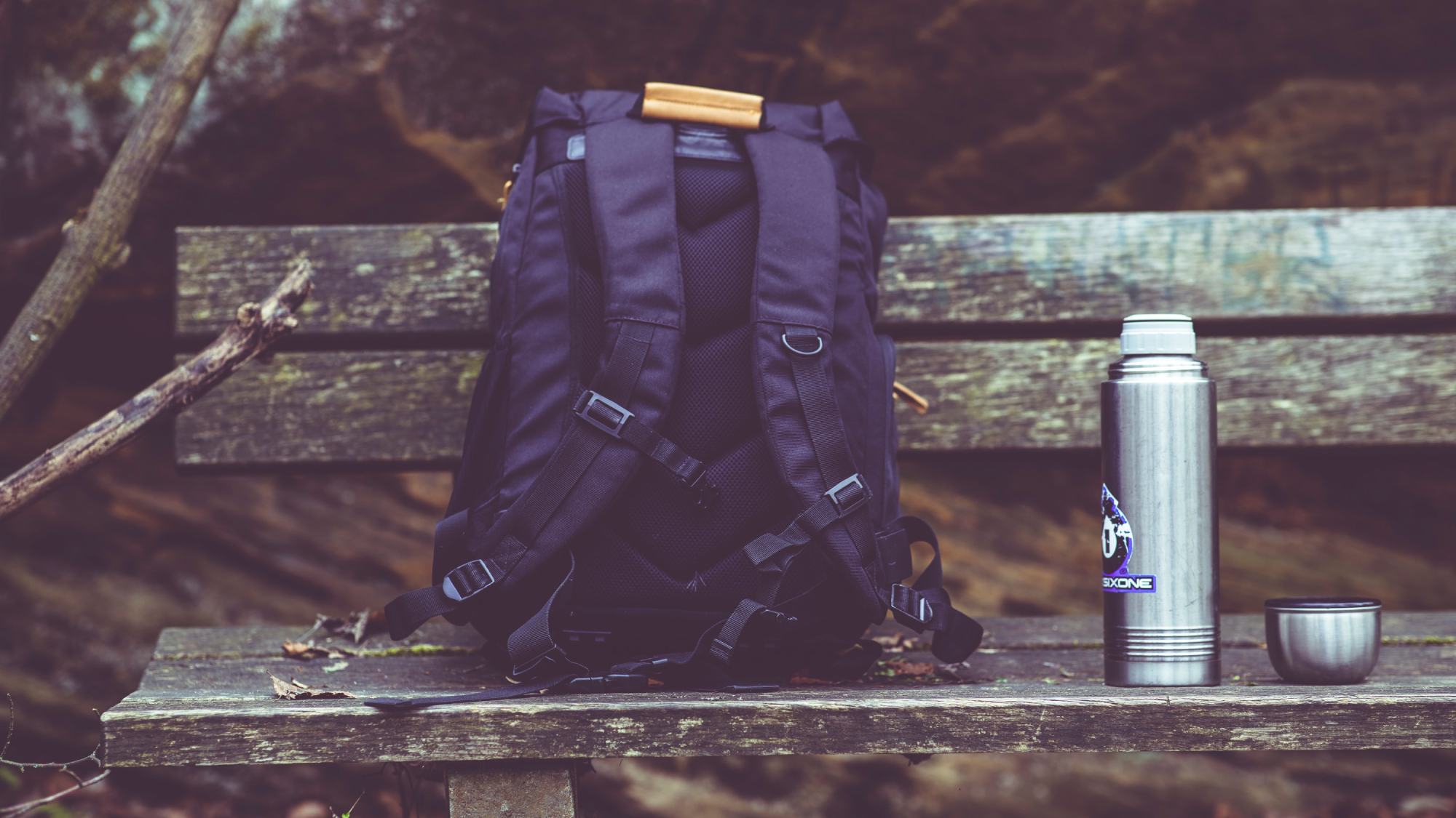 A black backpack on a weathered wooden bench in the forest, with a silver metal water bottle next to it.