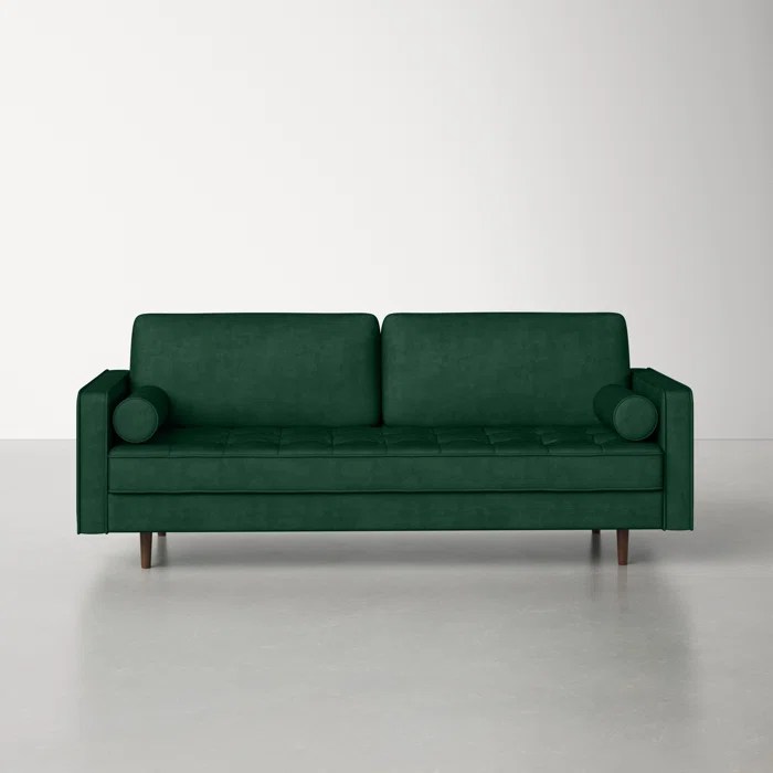 A green velvet couch on an off white floor and in front of an off white wall.