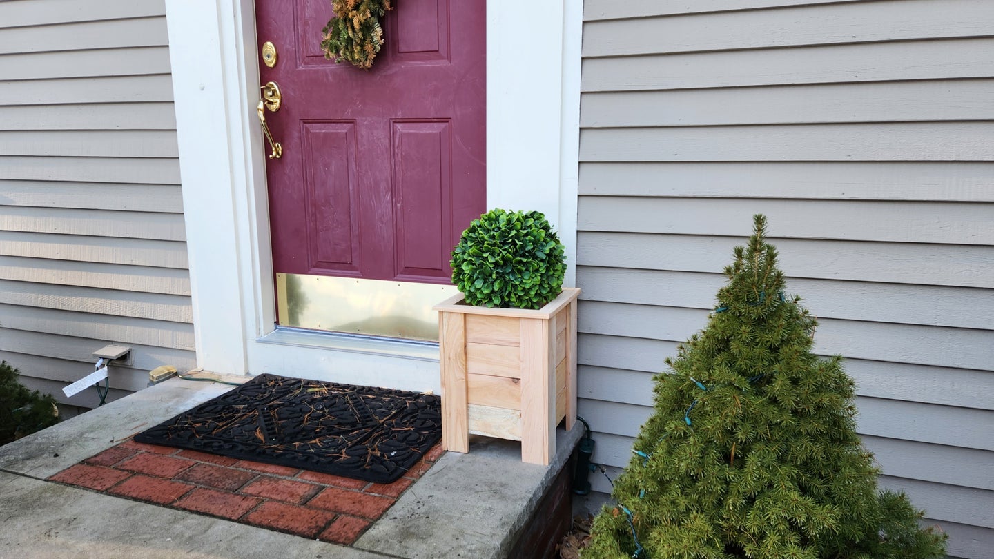 A cedar DIY planter box on the concrete front steps of a gray house with a red door.