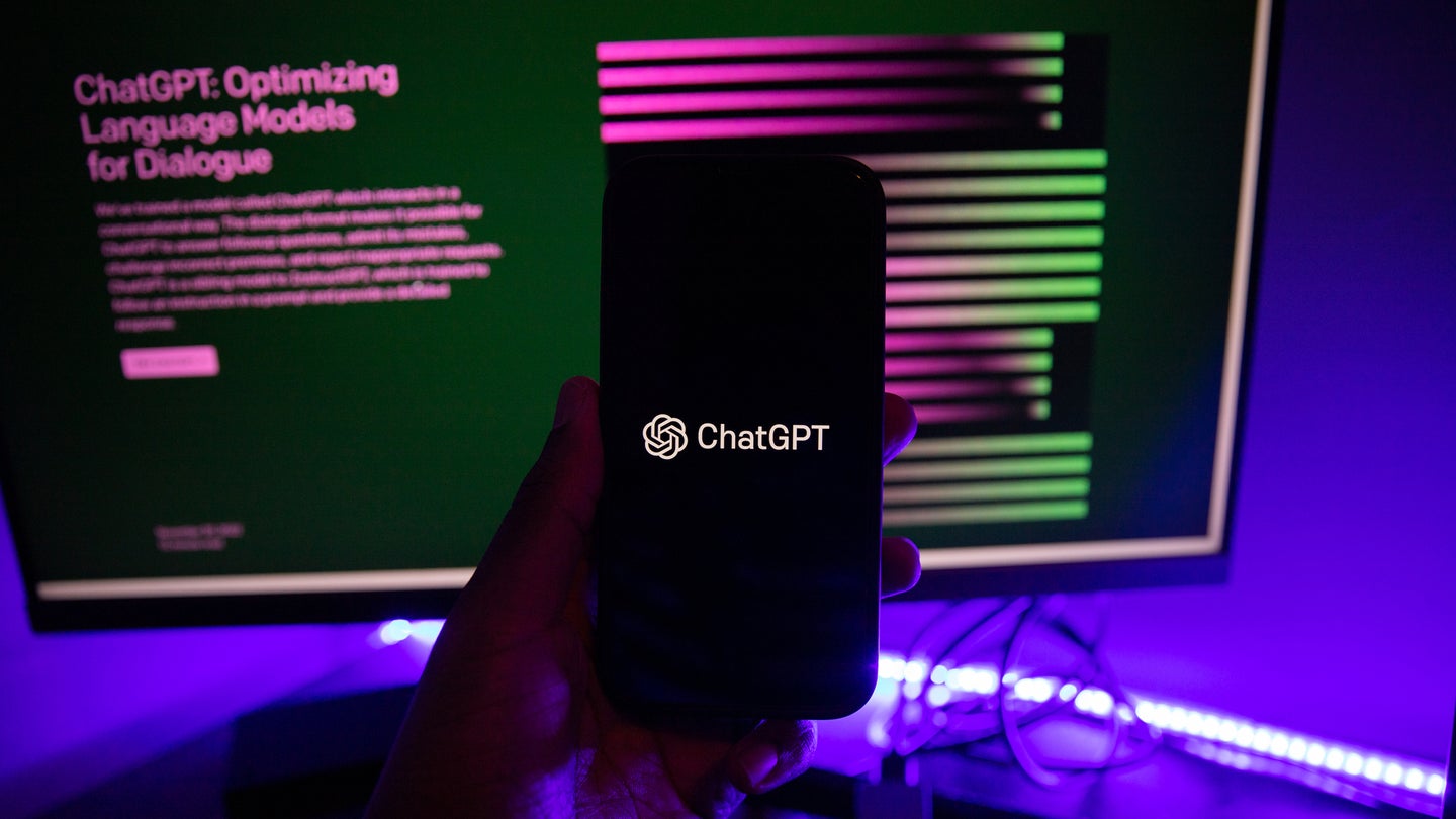 Screen of a smartphone with the ChatGPT logo in front of ChatGPT homepage on desktop monitor