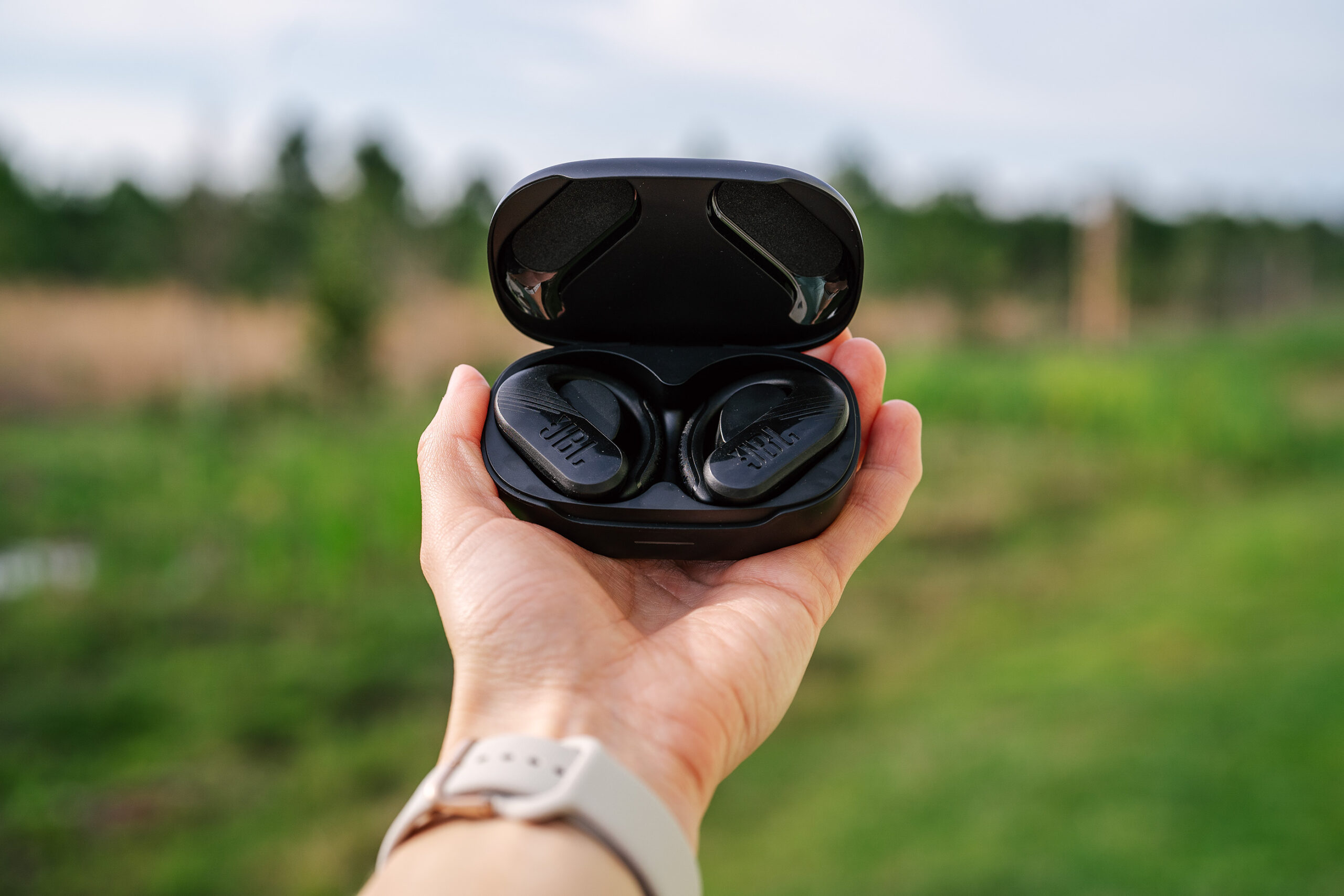 A person hold a pair of black JBL Endurance 3 headphones against a farm-like background.