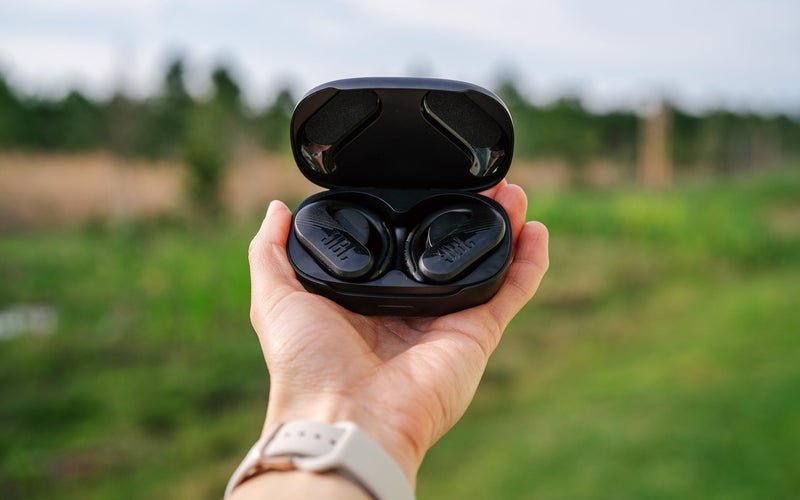 A person hold a pair of black JBL Endurance 3 headphones against a farm-like background.