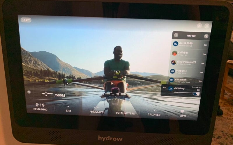 The touchscreen of the Hydrow Wave Rower, which features a world-class rower on a river with mountains in the background demonstrating how to row.