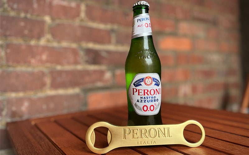 A bottle of Peroni NA on a wooden table in front of a brick wall.