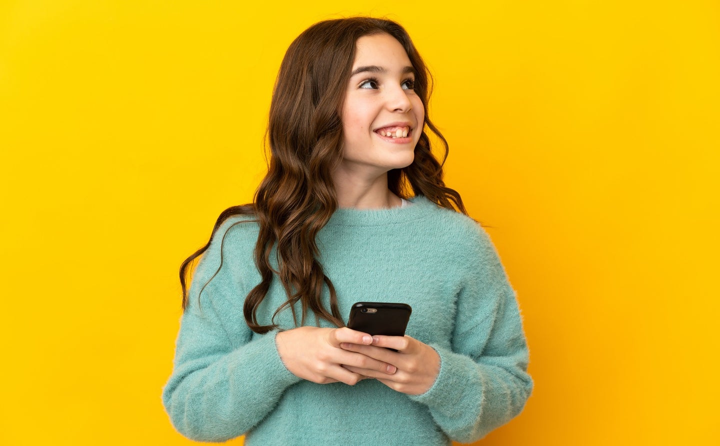 Teen in green sweatshirt with long brown hair against a bright yellow background scrolling through TikTok on a smartphone